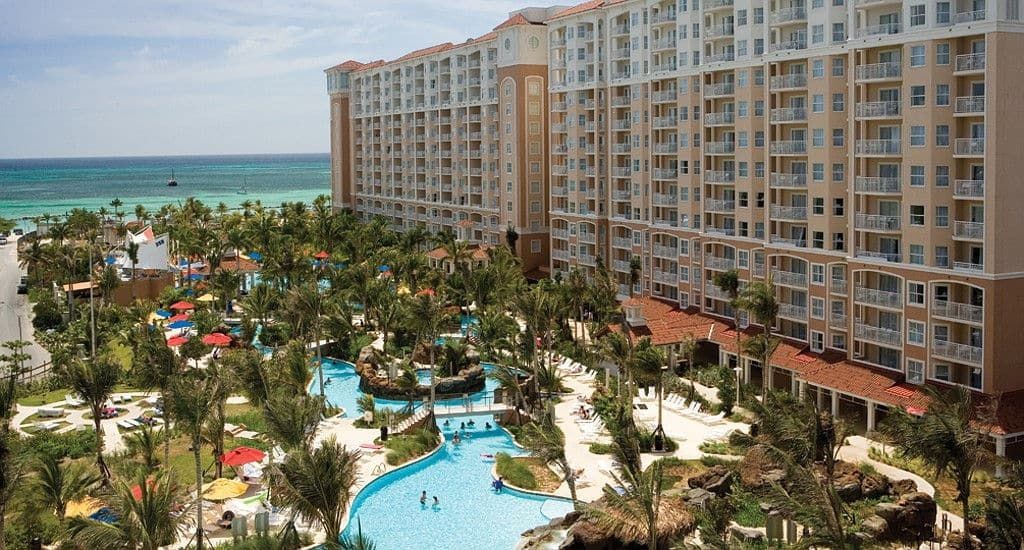 Large beachfront resort with multiple pools, palm trees, and lounge areas surrounded by high-rise buildings.