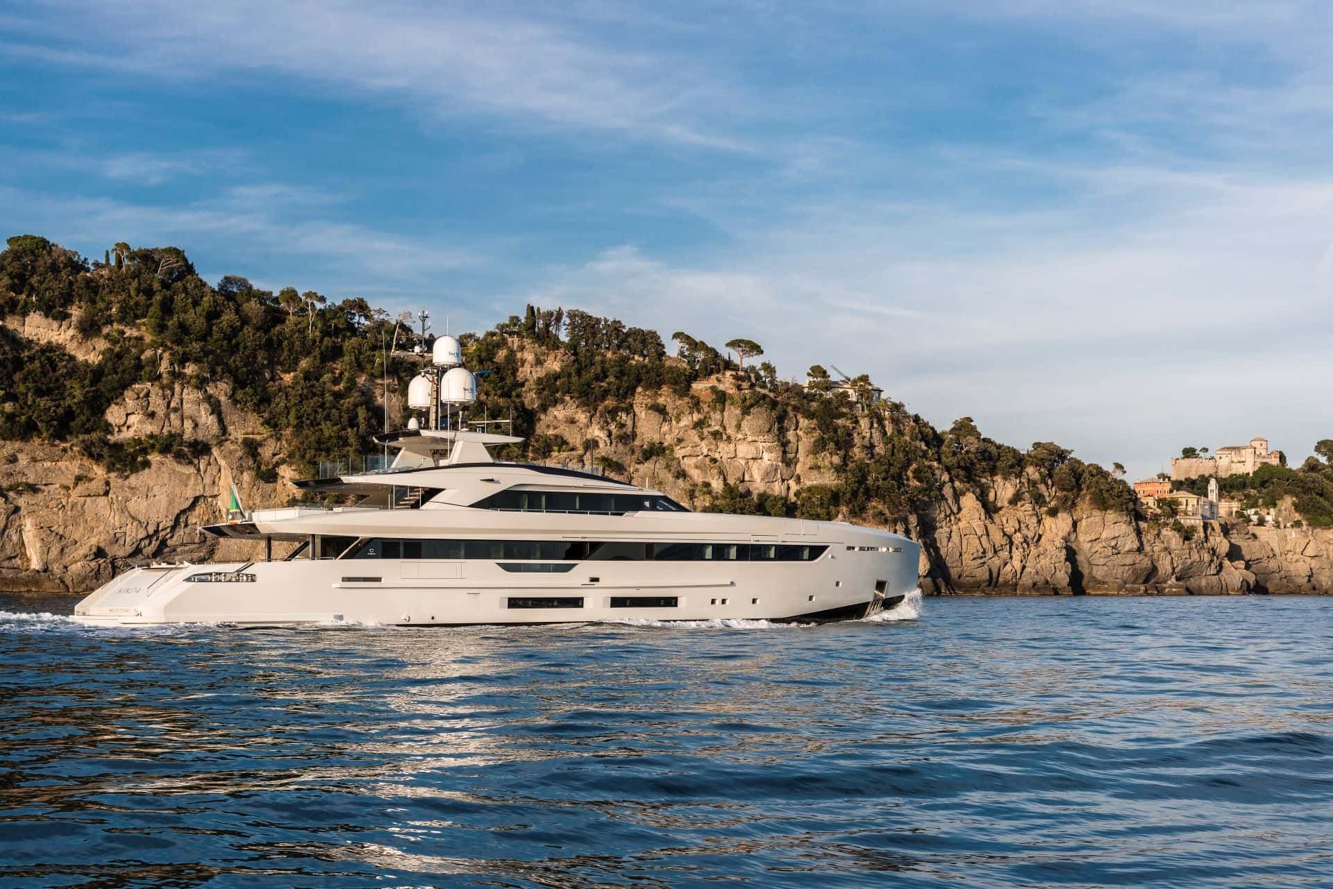 Luxury yacht sailing near a rocky coastline with trees and buildings under a blue sky.