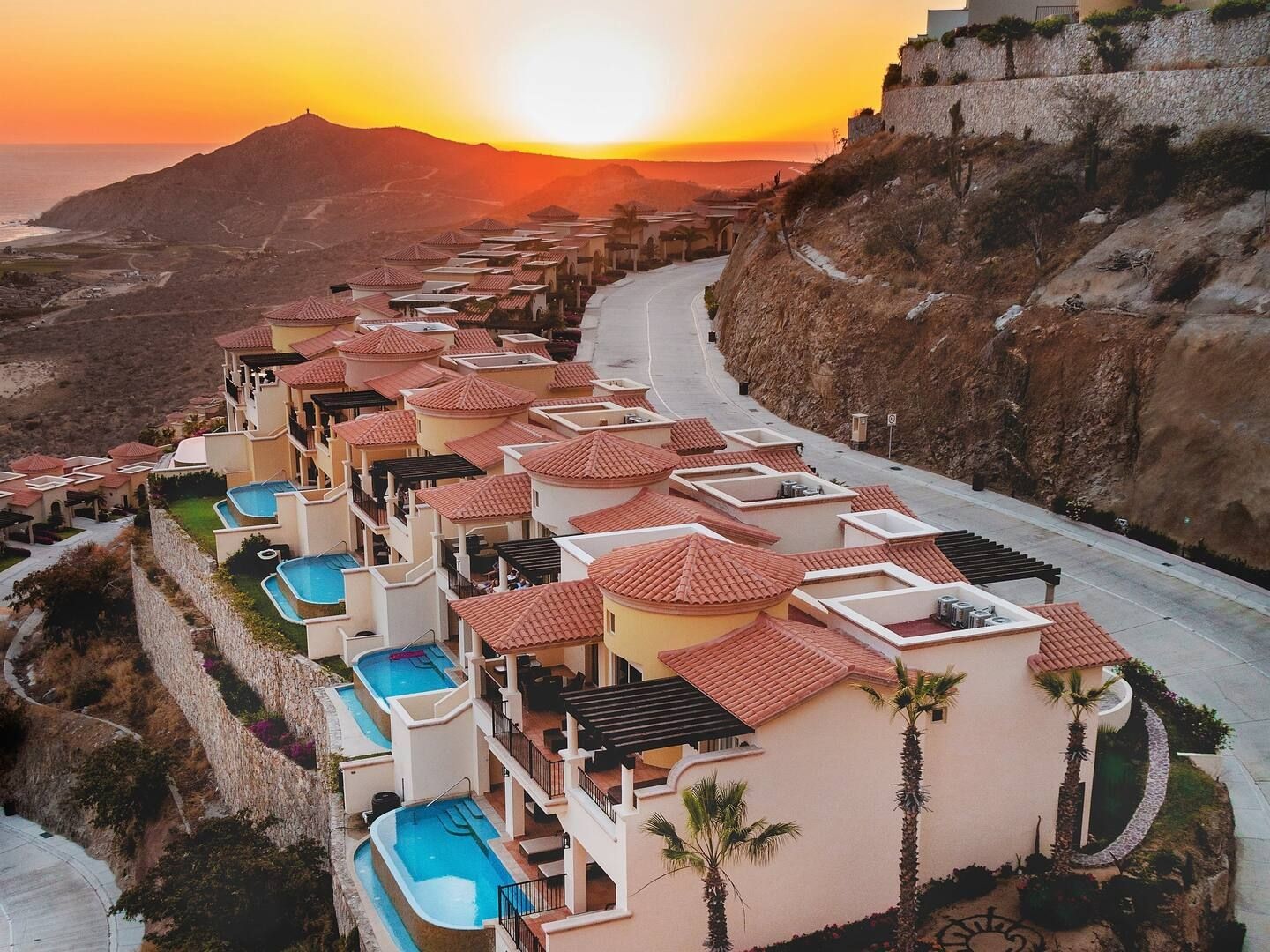 Aerial view of luxury villas with private pools on a hillside overlooking the ocean at sunset.