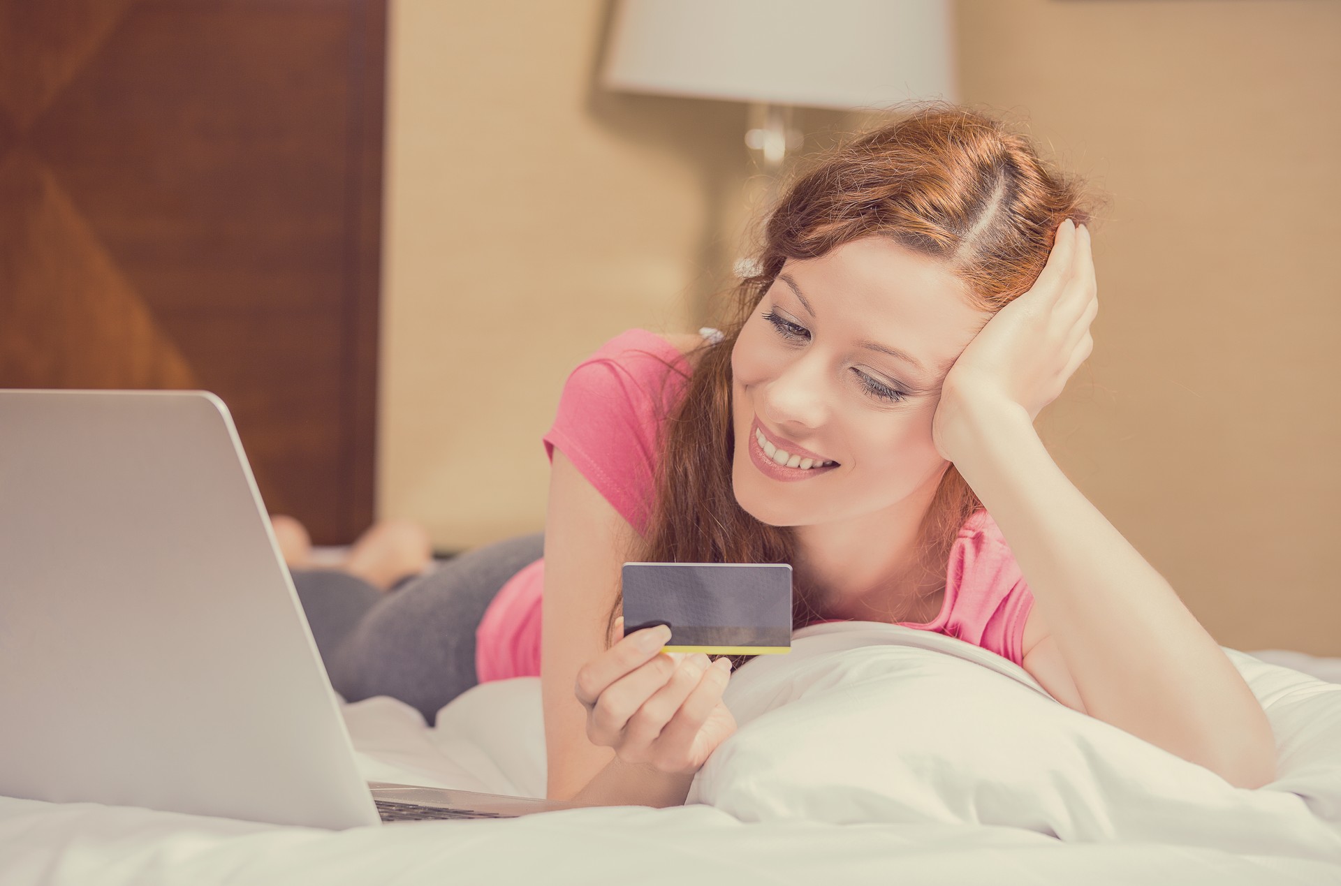 Cute young casual woman laying in bed smiling with laptop computer, holding credit card shopping online making reservation